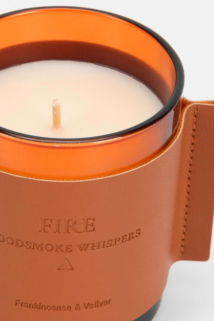 Candle in an orange holder with 'Fire Woodsmoke Whispers' text on a white background