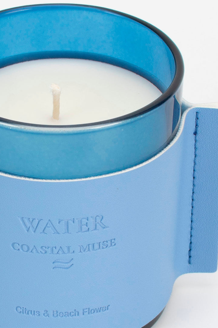 Candle in a blue holder with 'Coastal Muse' branding on a white background