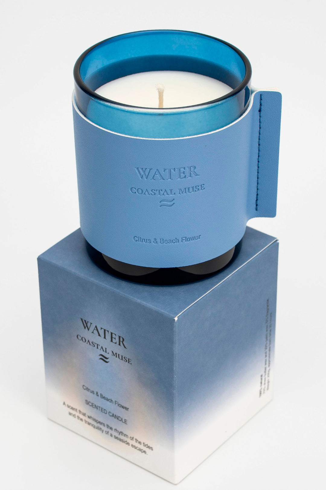 Candle in a blue container on a matching blue box with 'Water Coastal Muse' branding.
