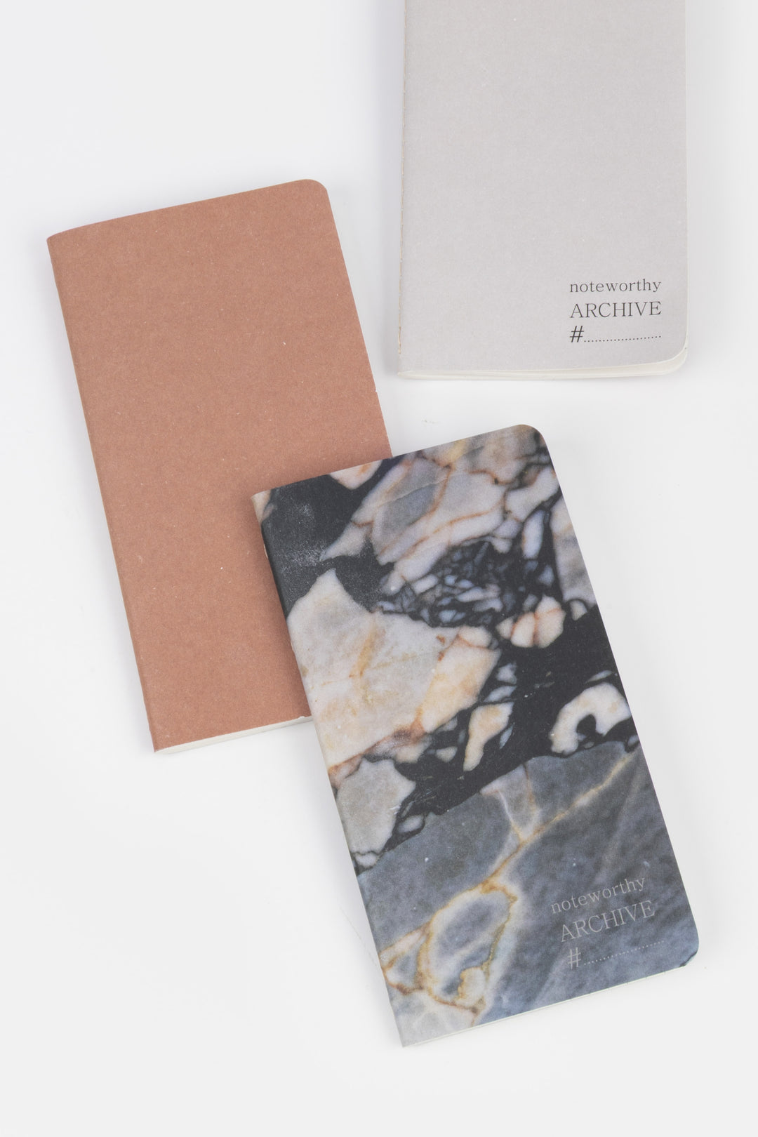 three mini softcover notebooks overlapping with one marbled cover one brown cover and one light grey cover
