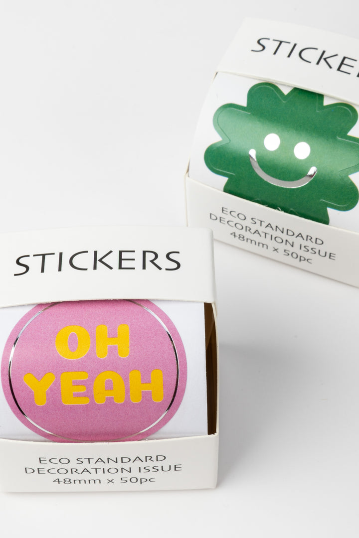 Packaged stickers with a pink one saying 'OH YEAH' and a green one with a smiley face on a white background.