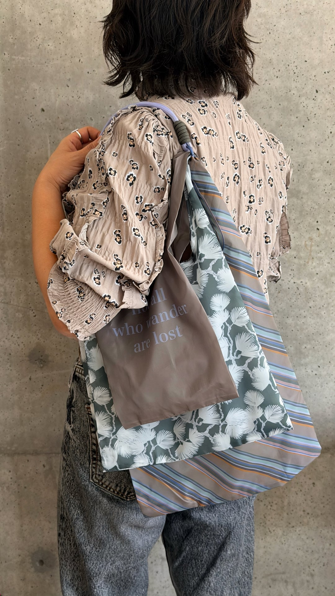 A person viewed from behind wearing a patterned blouse and gray pants, carrying a layered set of tote bags against a concrete wall. The bags include a taupe fabric with text, a green floral print, and a blue striped pattern.