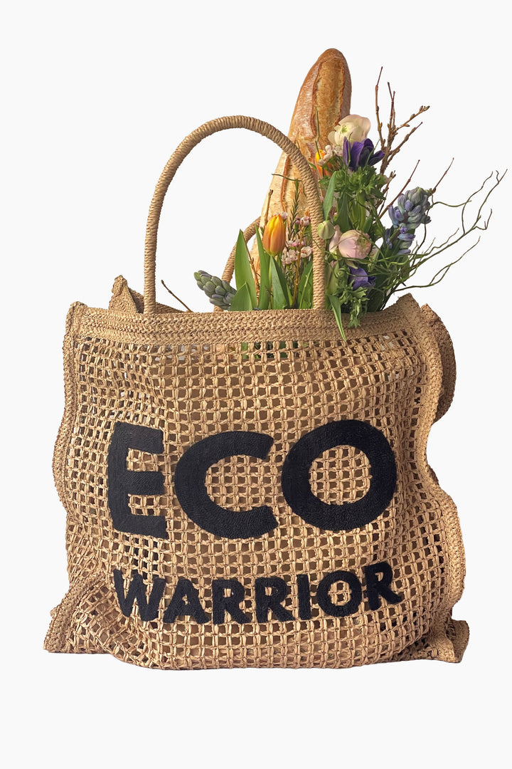woven paper straw bag with 'ECO WARRIOR' text filled with groceries on a white background